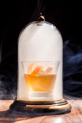 Photo of a smoked old fashioned under a domed container.