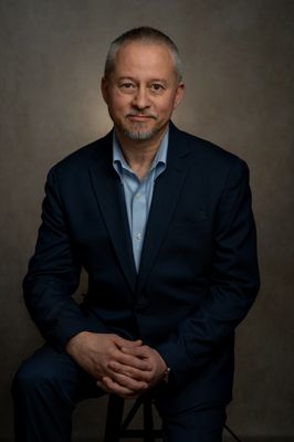 Confident mature man in navy suit, seated, hands clasped, radiating professionalism on a neutral backdrop