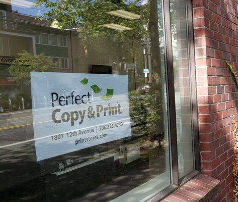 Come by our new location! DESCRIPTION: Perfect Copy & Print sign in window of retail store with brick façade.