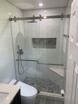 After. Floor to ceiling porcelain tile. Quartz nook, curb, and bench. Rain shower head with handheld. Frameless dual sliding glass doors.