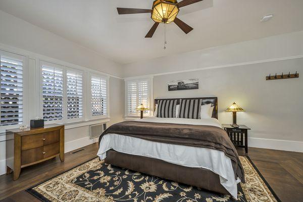 King bed with white bedsheets, brown throw blanket, and two striped pillows. Two side tables and lamps. Wall art and ceiling fan.