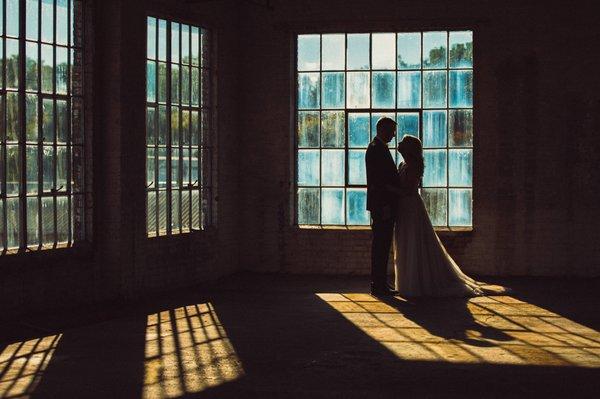 Couple embraces in front of windows in an industrial space. Photo by Ciao Bella Studios.