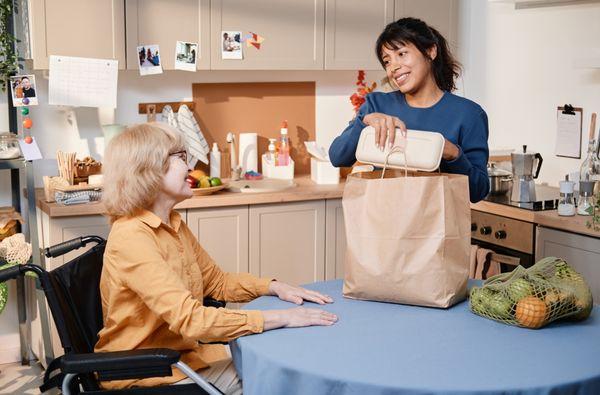 Smiling-Companions-in-caring-personnel-taking-food-out-paper-bag-she-delivering-food-for-senior-woman