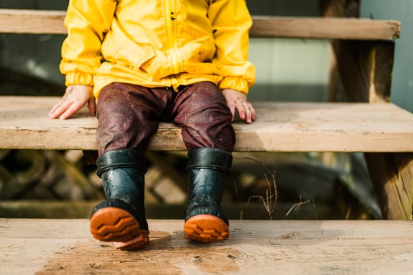 Toddler swings his legs while sitting on the porch steps wearing his muddy rain boots and rain coat