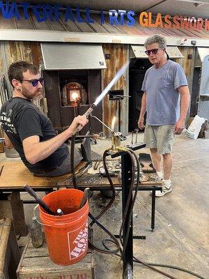 Waterfall Arts Board President, John Steed reaching for a glass blowing torch as Glassworks instructor David Jacobson looks on