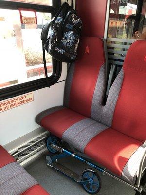 Kickscooter folded under seat, backpack hanging on corner knob. Seating configuration varies throughout the train; this is a roomier spot.