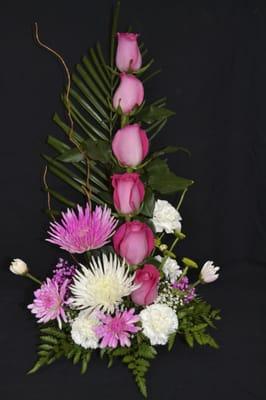 Floral arrangment lineal with 7 purple roses, 2 spiders, carnations pom poms, and green leaves.