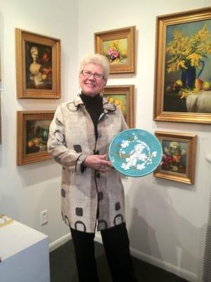 N. McElroy holding her ceramic plate and wearing a reversible art jacket  sold at the gallery..oils by M. Duchesne