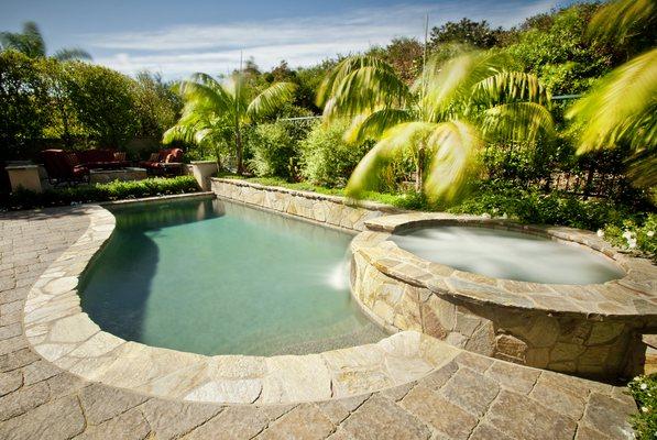 This tropical pool and spa design by Artisan Outdoor features custom stonework and a lush planting plan.