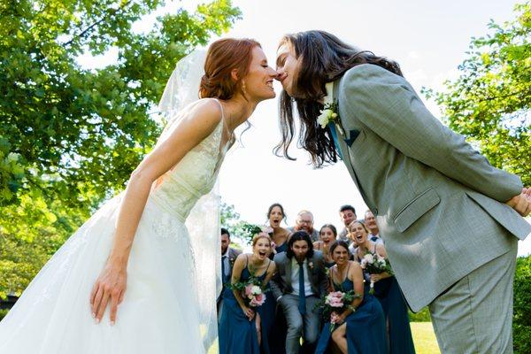 Wedding photo of couple kissing while wedding party looks on.