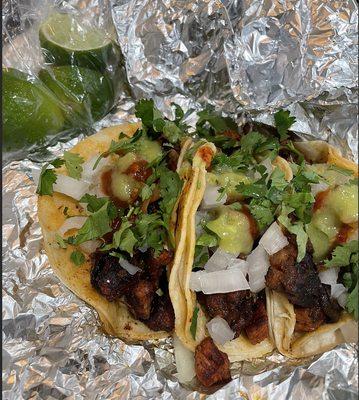 Three tacos al pastor with cilantro, onion, limes, and various sauces.