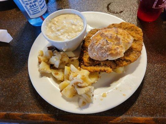 Country fried steak on a biscuit with a side of home fries and gravy.