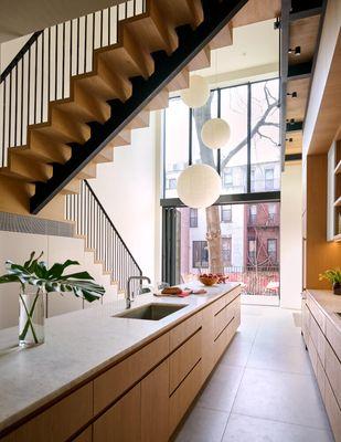 The glass wall behind the double-height kitchen fully opens to the garden beyond.  A stair dynamically climbs to the catwalk above.