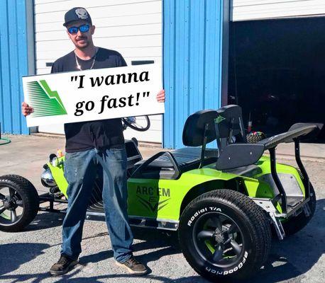 owner of arcem ev holding sign that says I wanna go fast. Standing next to a green rat rod racing golf cart