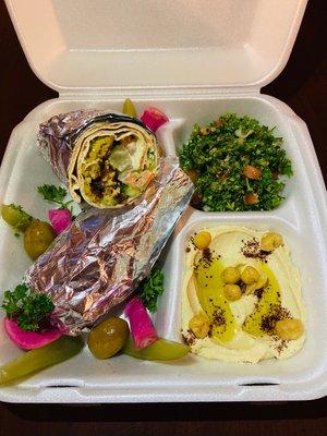 Falafel sandwich wrap, filled with tomato, cucumber, pickles, and drizzled with tahini sauce. Sides are tabouli salad, and hummus.