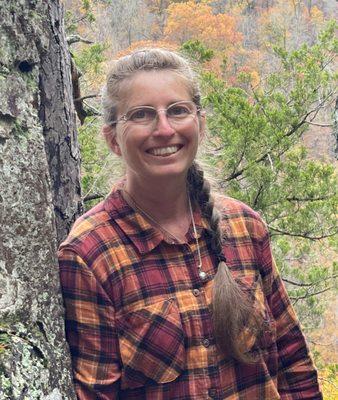 Woman in orange/red plaid smiling leaning against tree with braids