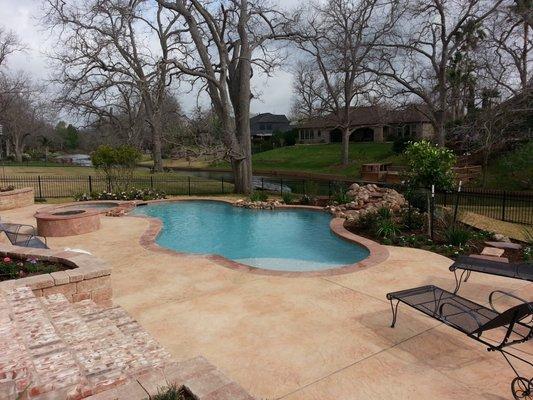 Pool with leveled spa, waterfall, travertine deck overlaid on concrete. Retention wall with steps, and gas fire pit.