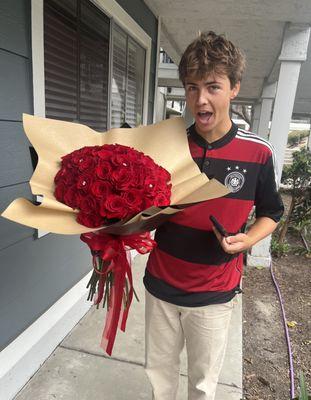 A dashing young man stands tall, clutching a vibrant bouquet of velvety roses. The rich crimson petals complement his stylish attire.