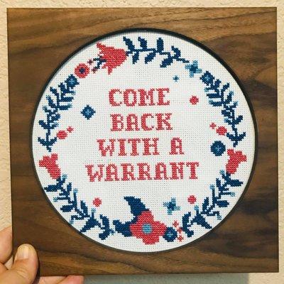 "Come back with a warrant" on cross-stitch art piece.