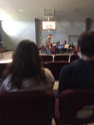 Pastor Ben teaching. Seats are in a semicircle in a carpeted gymnasium.