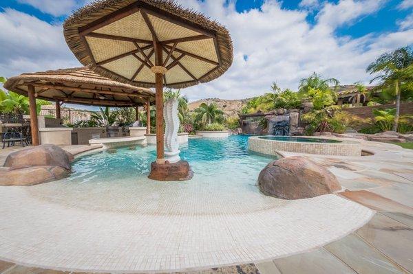 Tropical beach entry custom swimming pool with a perimeter flow spa, grotto, rock slide, and waterfalls. Outdoor kitchen with swim up bar.