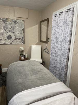 Lovely treatment room with gray and white beautiful floral picture and curtain with facial bed with cozy gray blanket.