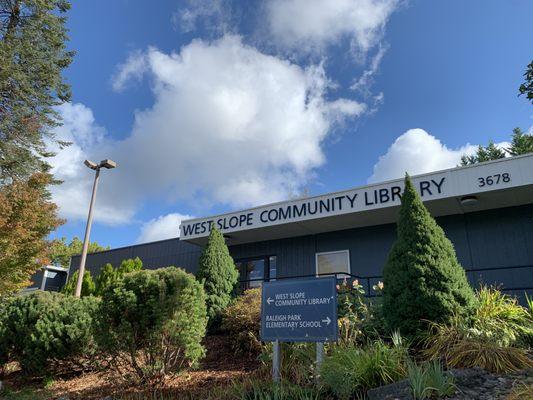 Front of West Slope Community Library building in spring, with a lot of fluffy white clouds overhead.