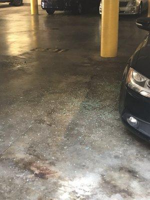 Broken glass all over the garage apartment complex floor & spilling into the side of the adjacent parking spot.