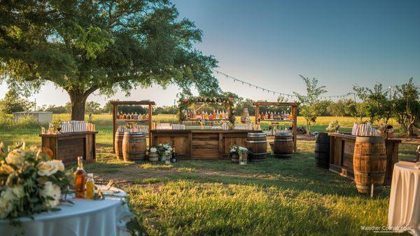 A charming rustic bar setup with wooden stations and barrels under tree lights, perfect for weddings and casual gatherings.