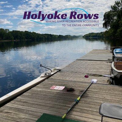 A single scull boat sits ready at the on the Connecticut River. The words "Holyoke Rows" appear across the top