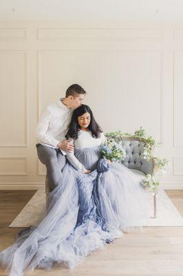 A man and woman sit in an elegant chair while admiring their baby soon to come. they look elegant and dreamy. The blue tulle skirt flows