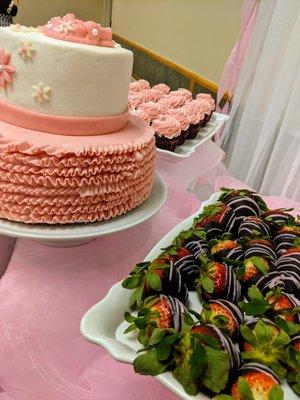 A baby shower dessert table with a custom layer cake, cupcakes, and chocolate covered strawberries.