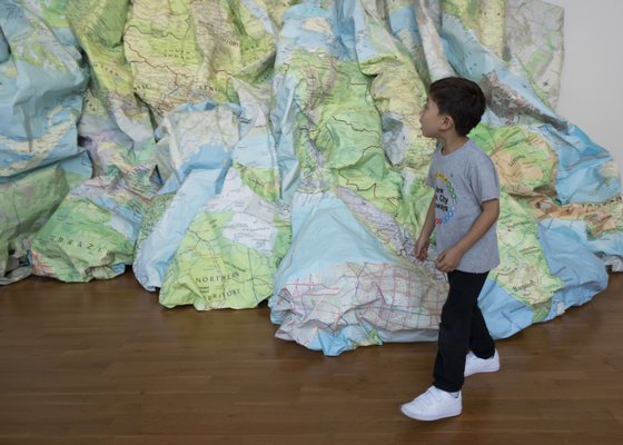 Boy standing in front of large sculpture made our of crumpled maps.