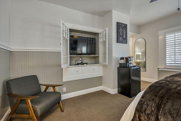 King bed with white bedsheets and brown throw blanket. Black and white wall art. Black leather armchair. Flatscreen TV in cupboard.