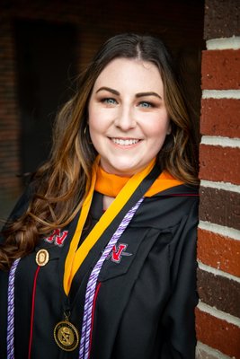 Masters graduate smiles from behind a brick column wearing her regalia.