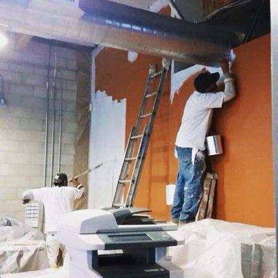 The workers are paint rolling and hand brushing the wall.