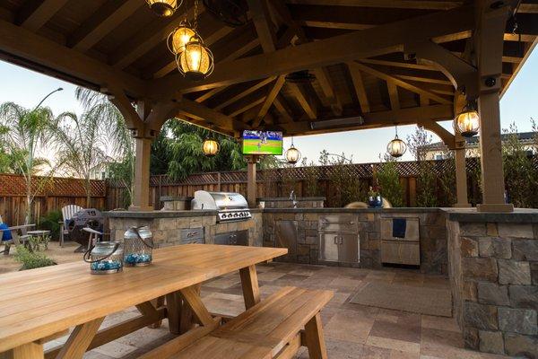 Picnic bench with outdoor kitchen, custom pergola, flatscreen TV, BBQ, and custom lighting.