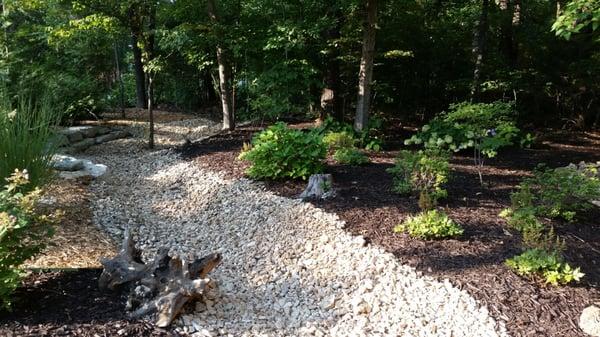 Installation of a dry creek helps contain runoff water from the slope above.  Surrounding plantings and mulch add beauty to t...