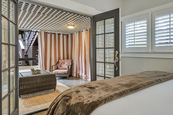 King bed with white bedsheets and brown throw blanket. Door with shutters on window leading to private patio.