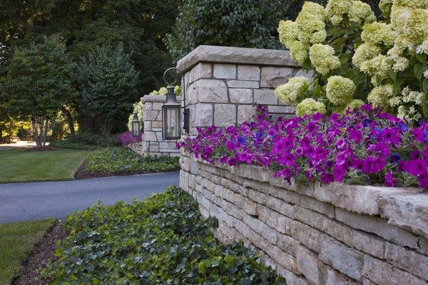 Welcome home to our grand entrance design featuring stately pillars with luminous lamps, flanked by lush greenery and vibrant flowers.