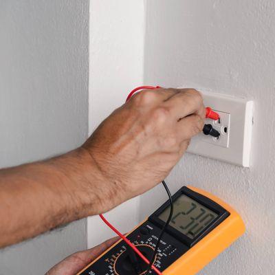 Inspector using a digital meter to measure the voltage at the power outlet in on the wall.