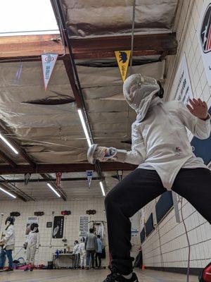 Me, posing for a selfie in my gear, with others fencing in the background.