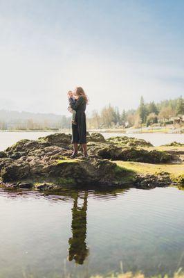 A mother kisses her son on a lake with their reflection. You can see the sky and horizon with the lake and forest in the background.