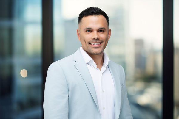 A man in a light blue blazer and white shirt smiles warmly for a headshot , set against a blurred cityscape background with large windows.