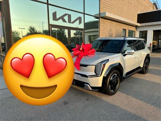 Heart eyes emoji next to an ivory white Kia EV9 with a red bow on the hood. Kia dealership in background.