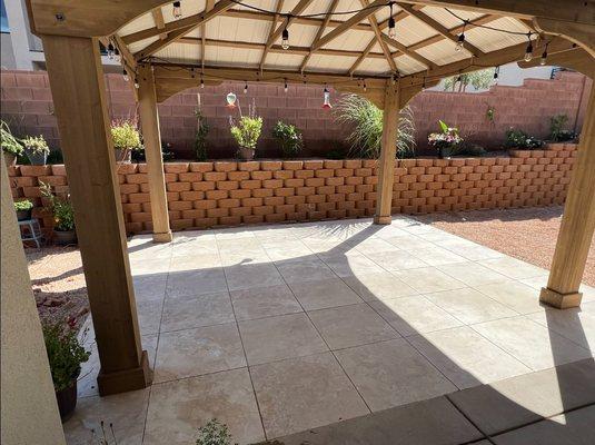 A covered patio area with porcelain pavers on the floor. The patio is supported by wooden posts and beams, and it has a roof providing shade