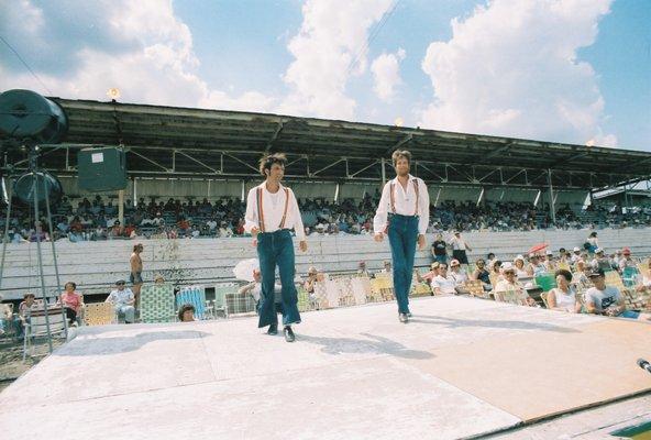 View of the grandstands at Clark County Fairgrounds, with open air seating area in front, and Tom and Mark completing their buck dance.